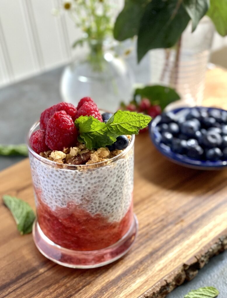 Chia pudding in a glass topped with berries and mint leaves on top of a wooden slab with a bowl of blueberries in the background.