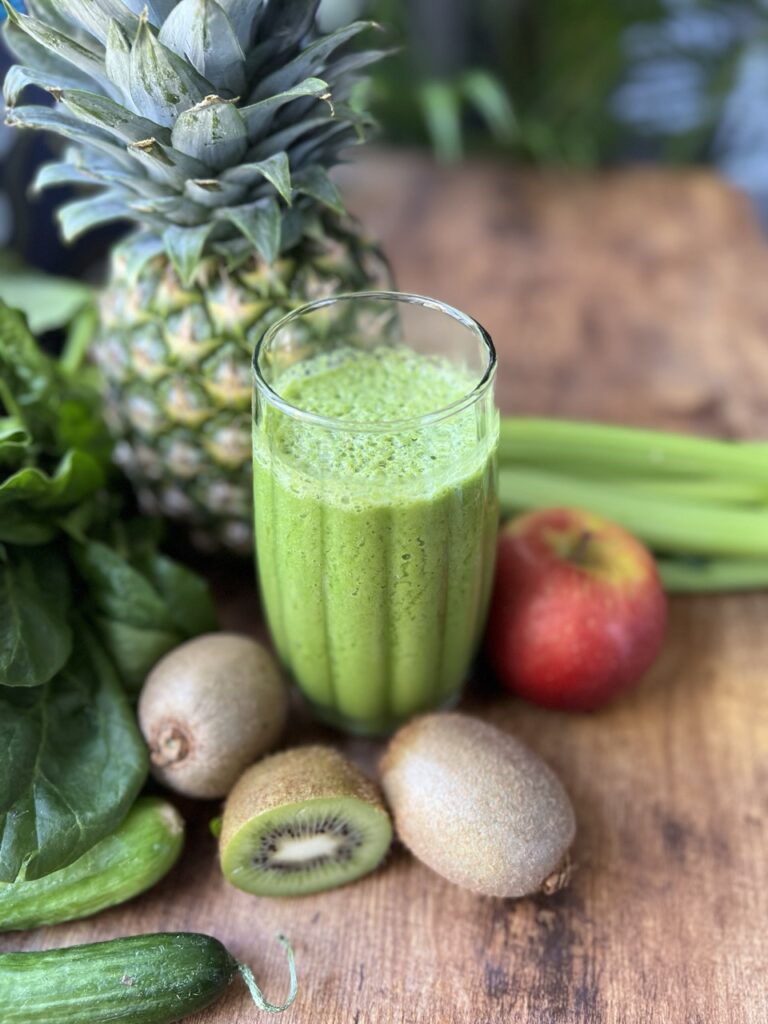A glass of green smoothie on a table surrounded by the fruits and vegetables that it's made of:  kiwi, cucumber, celery, apple and pineapple.