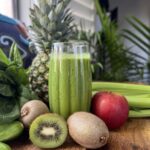 Eye level view of smoothie in front of a window surrounded by the fruit and vegetables that comprise it: kiwis, apples, celery, cucumbers, spinach and a whole pineapple.