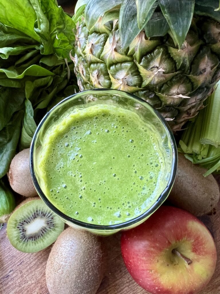 Overhead image of a green smoothie surrounded by the fruit it's made from:  apples, kiwis and pineapple as well as spinach.