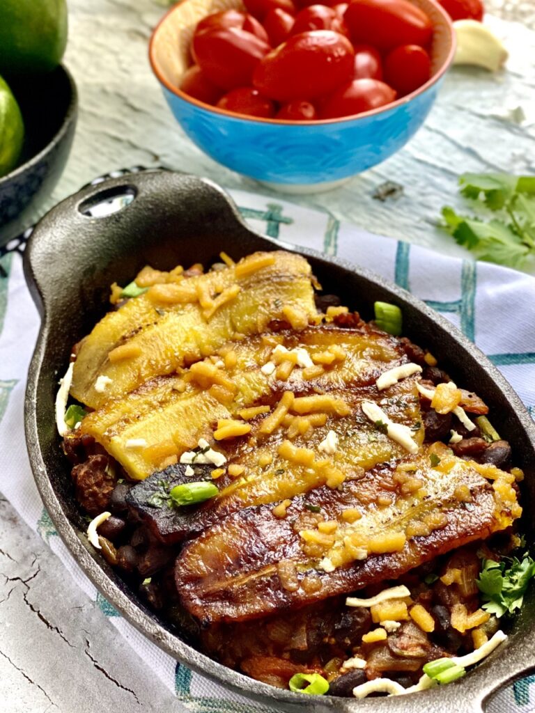 Pastelon in an oval cast-iron casserole dish on a table with a bowl of tomatoes in the background.