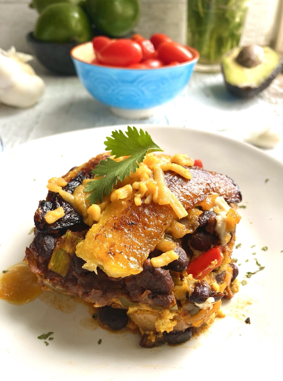 Slice of pastelon on a plate with a bowl of tomatoes and a halved avocado in the background