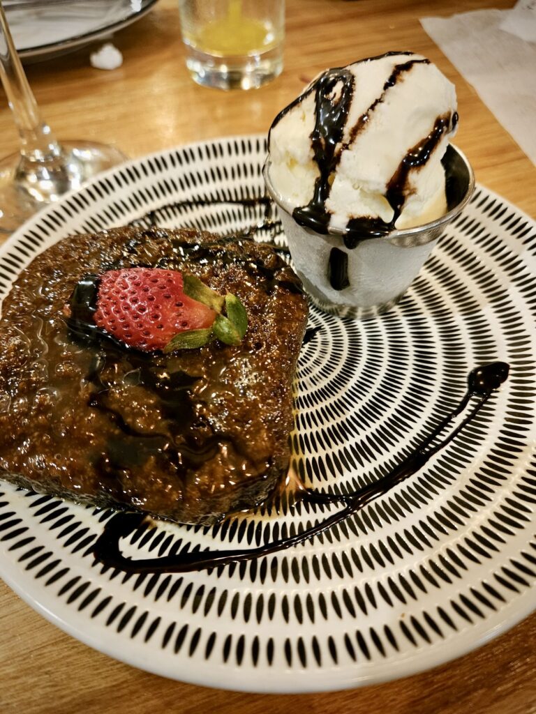 A plate with a piece of malva pudding topped with a single strawberry and a drizzle of chocolate, with a cup of ice cream on the side.