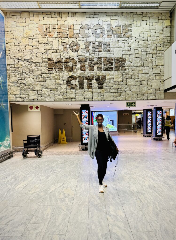 Photo of woman standing in front of  of stone mural with words welcome to the mother city created by stones