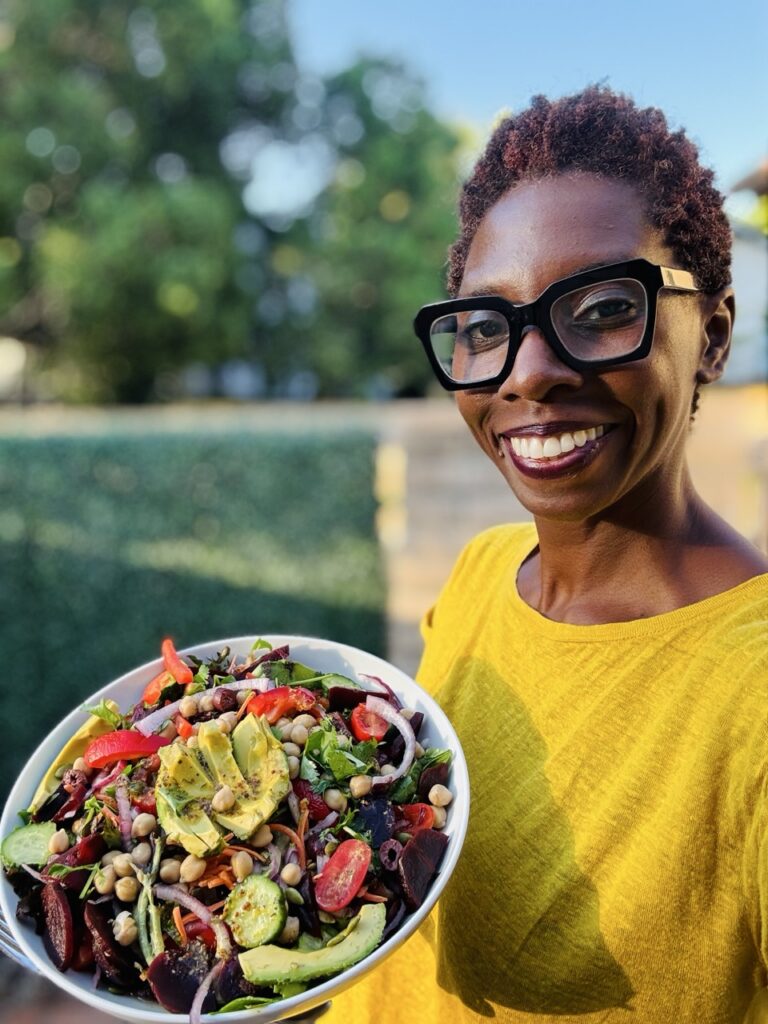 Picture of woman in yellow shirt and glasses holding a bowl of Everything said with field greens, arugula, shredded carrots, bell peppers, red onions, avocado, dates, beets, chickpeas, pumpkin seeds, and tomatoes.