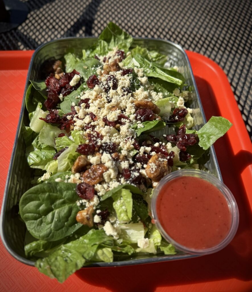 Large salad in a metal tray topped with candied pecans, cranberries and feta served with a side of raspberry dressing.