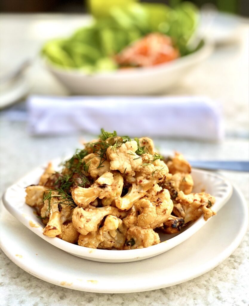 Bowl of charred cauliflower topped with pistachios, chopped dates and dill with green salad in the background.