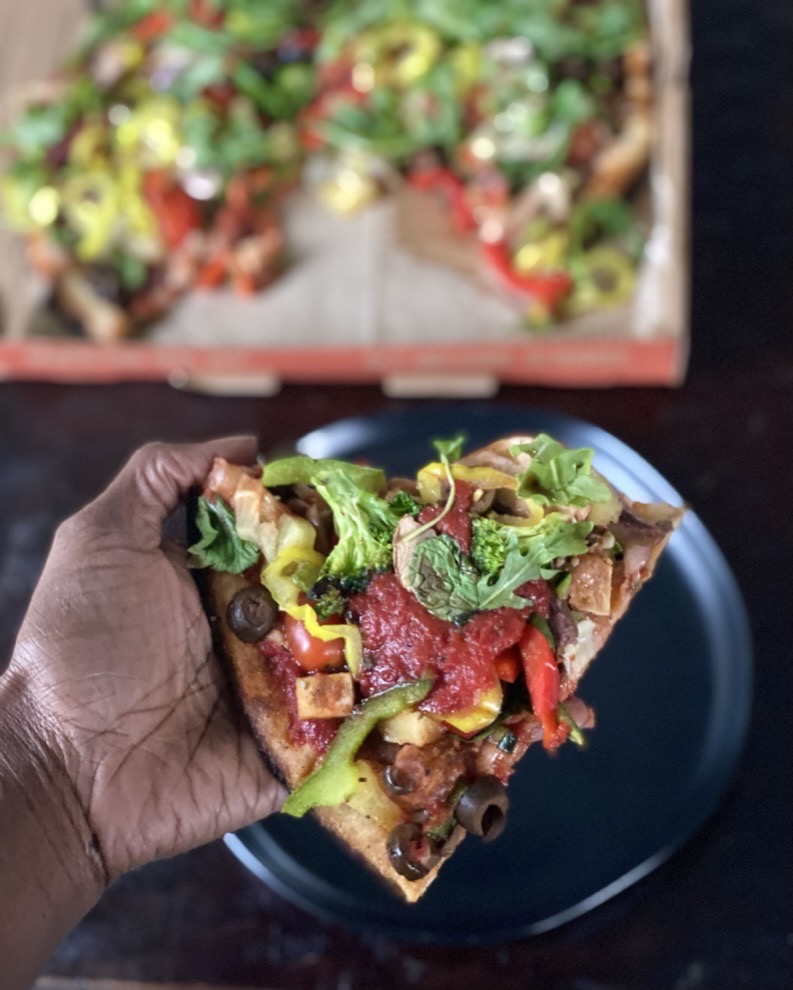 Slice of Blaze pizza loaded with veggies being held over a plate with a box of pizza in the background.