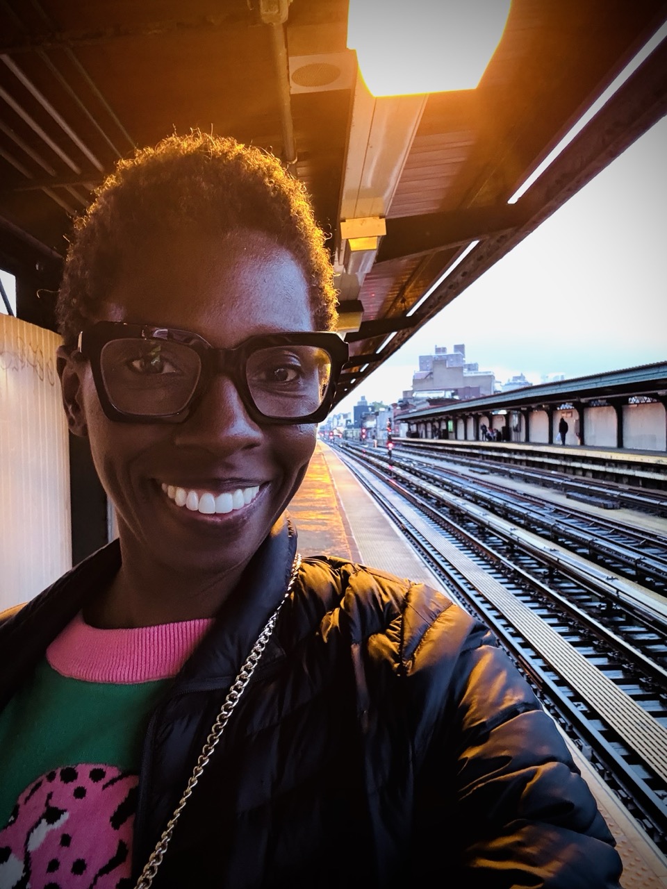 Smiling woman wearing glasses standing on a subway platform at sunset.