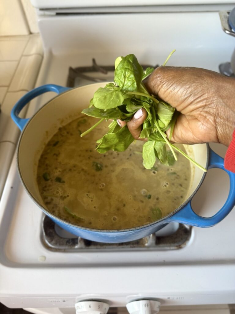 A pot on a stove filled with mung bean soup with a hand filled with spinach over the pot.
