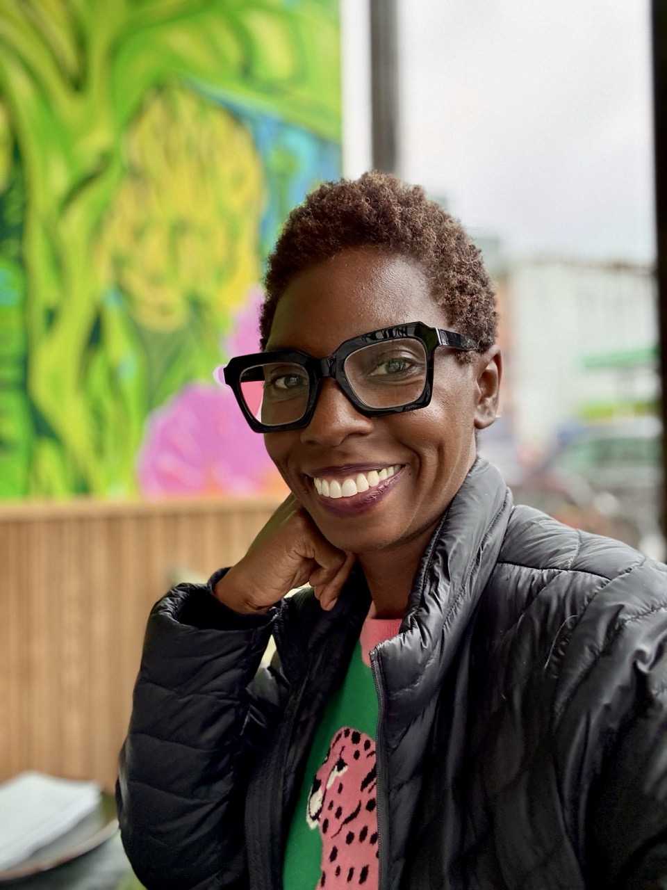 Smiling woman wearing glasses sitting at a table with her hand on her chin.