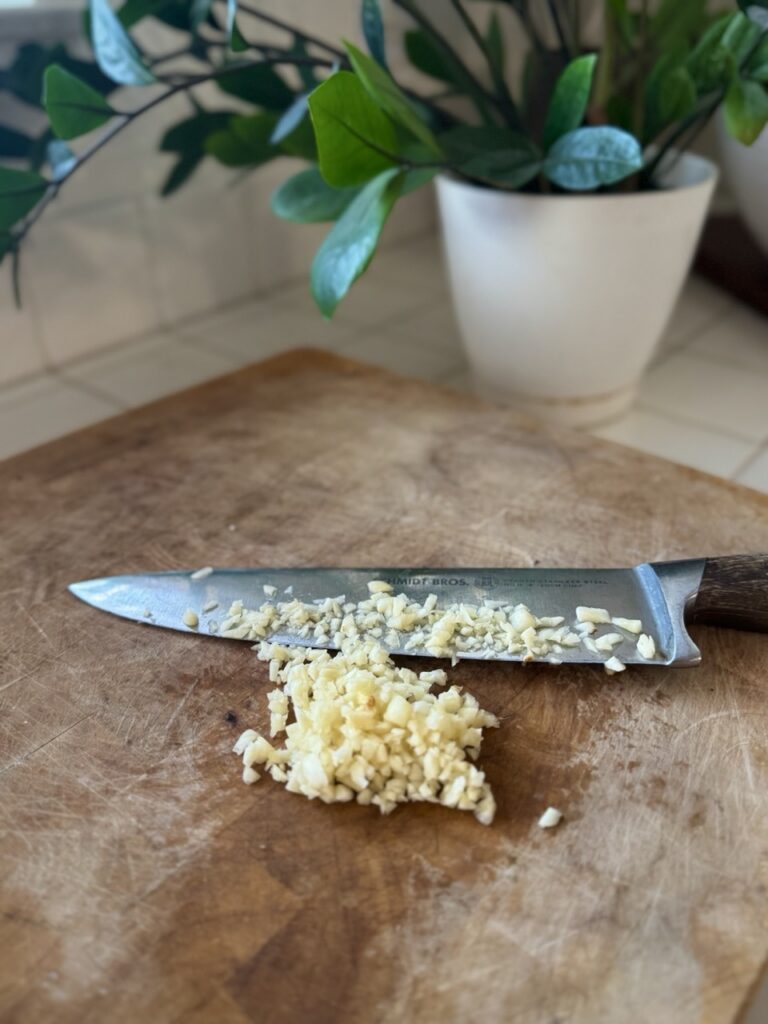 Diced garlic with a knife on a cutting board with a green z plant in the background.  