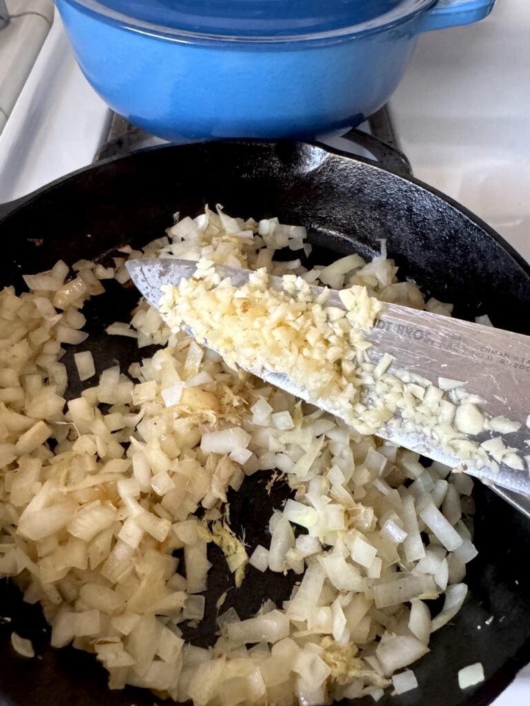 A cast iron skillet on the stove filled with mined garlic, mined ginger and diced onions with a knife held over the skillet.
