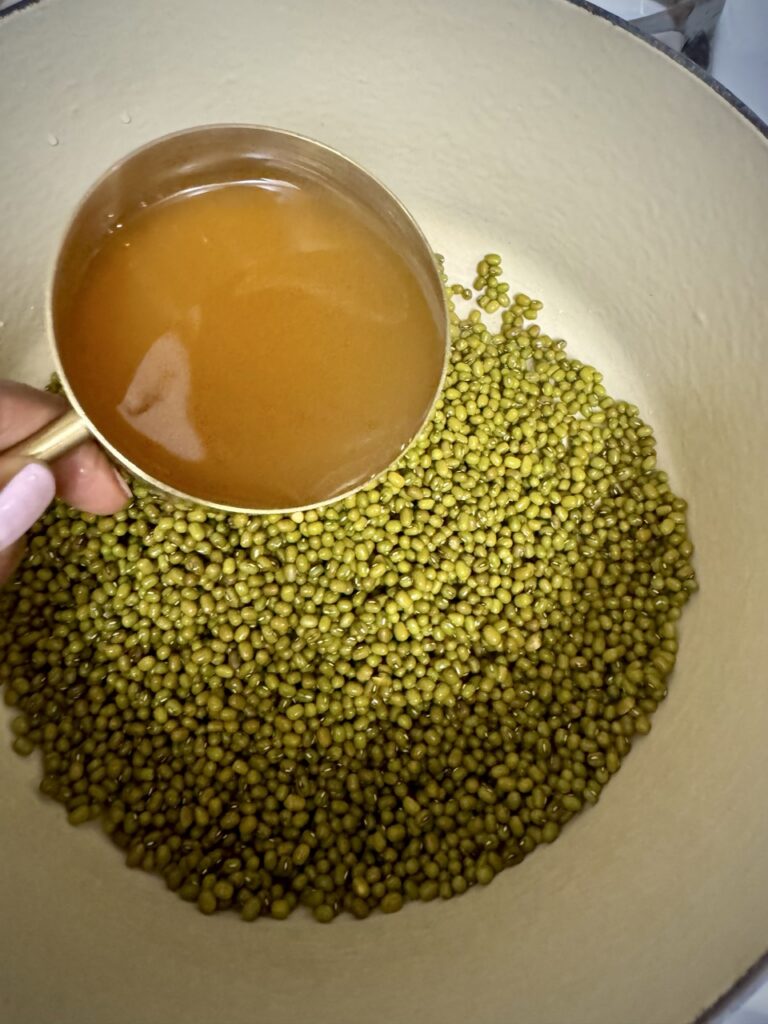 Dried mung beans in a pot with a measuring cup of vegetable broth about to be poured over it.