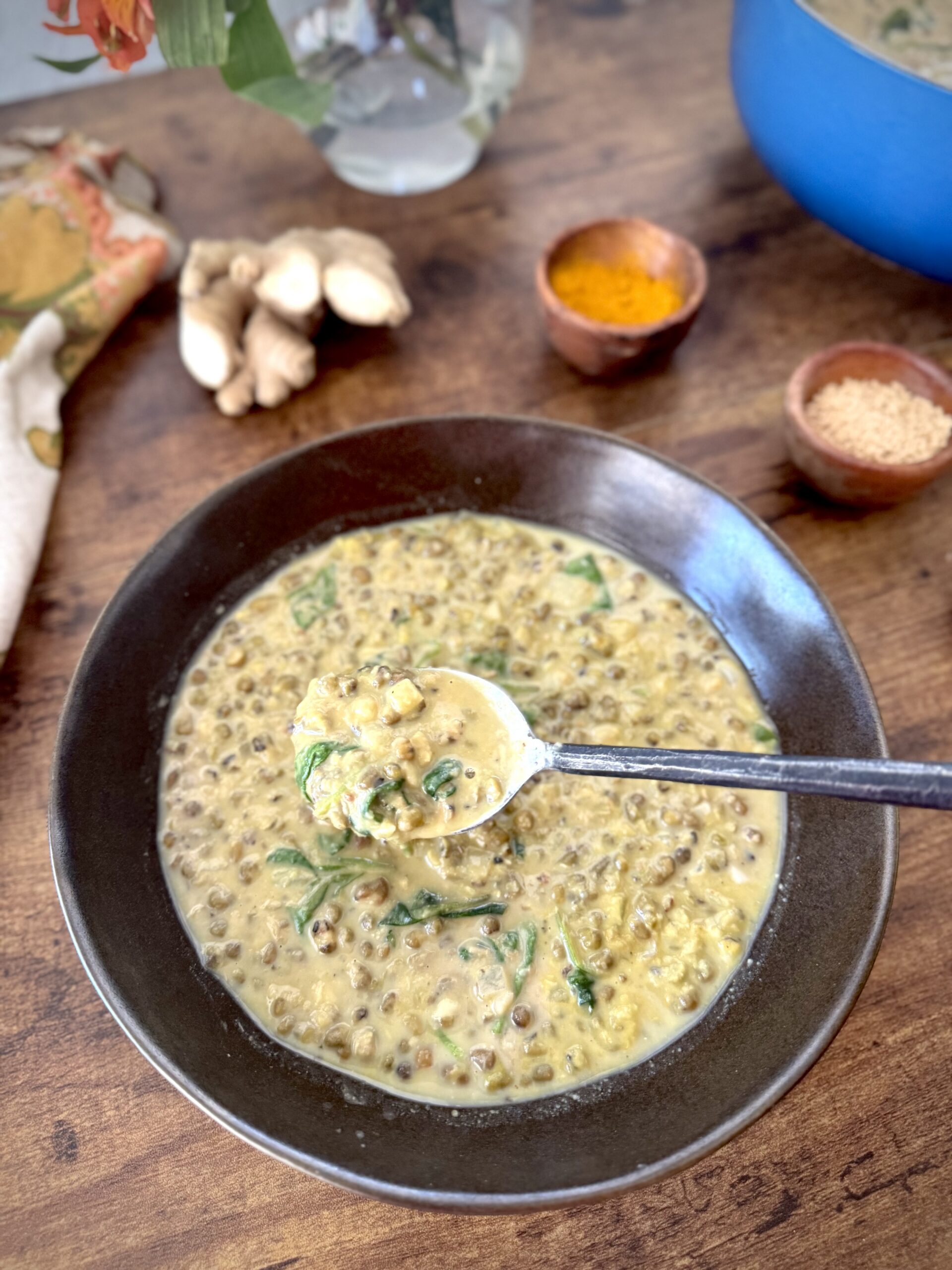 Bowl of mung bean soup with spoon in it and fresh ginger and ground turmeric and mushroom powder in the background.