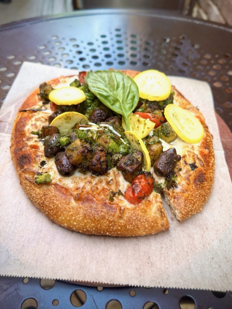 Urth Caffe's Pizza Primavera made with pesto marinated asparagus, potatoes, yellow squash, sugar plum tomatoes, Kalamata olives and Treviso lettuce. Baked on a vegan pizza crust with vegan Monterey jack cheese and topped with grilled artichoke hearts, basil flower and peperonata sauce.