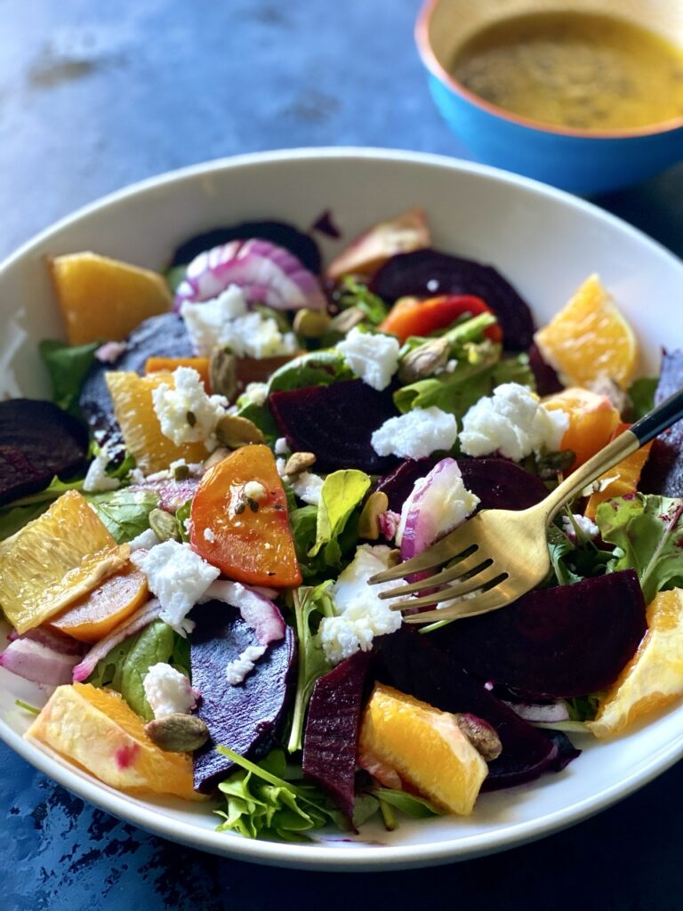Bowl of roasted beet salad with golden and red beets, arugula, red onion, orange segments and vegan feta.
