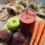 A glass of ABC juice with a metal straw in it, on a wooden platter with one green apple, two beets, and three carrots on it.