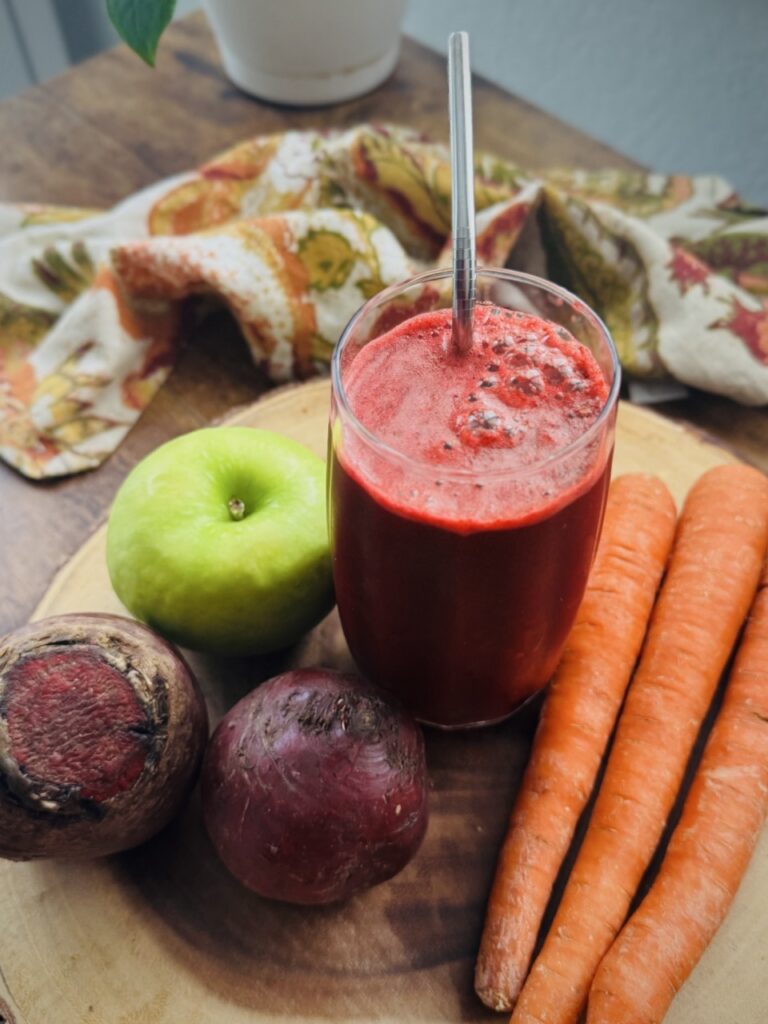 A glass of ABC juice with a metal straw in it, on a wooden platter with one green apple, two beets, and three carrots on it.