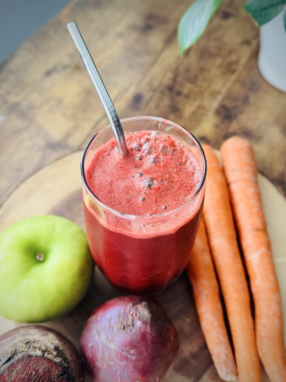 A glass of ABC juice with a metal straw in it, on a wooden platter with one green apple, two beets, and three carrots on it.