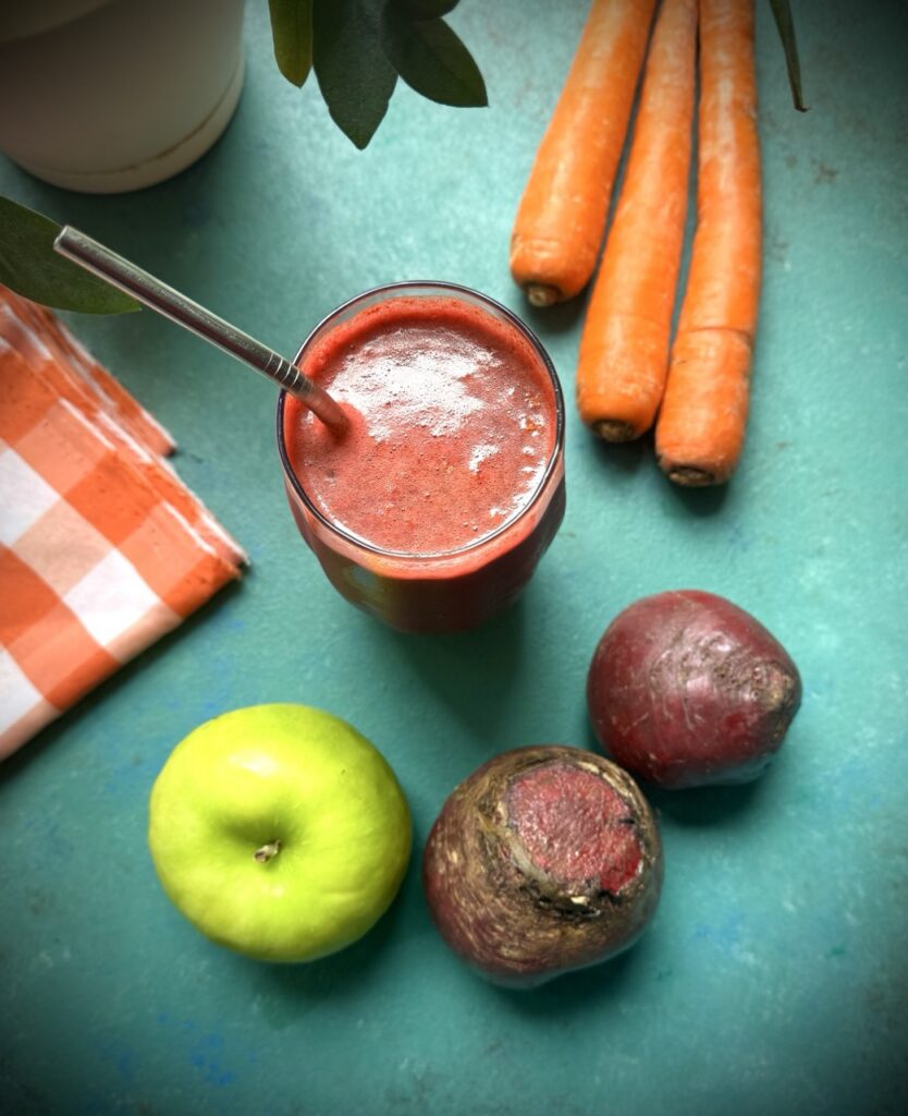 An overhead image of a glass of ABC juice with a metal straw in it, on a green backdrop with one green apple, two beets, three carrots and an orange and white checkered piece of fabric on it.