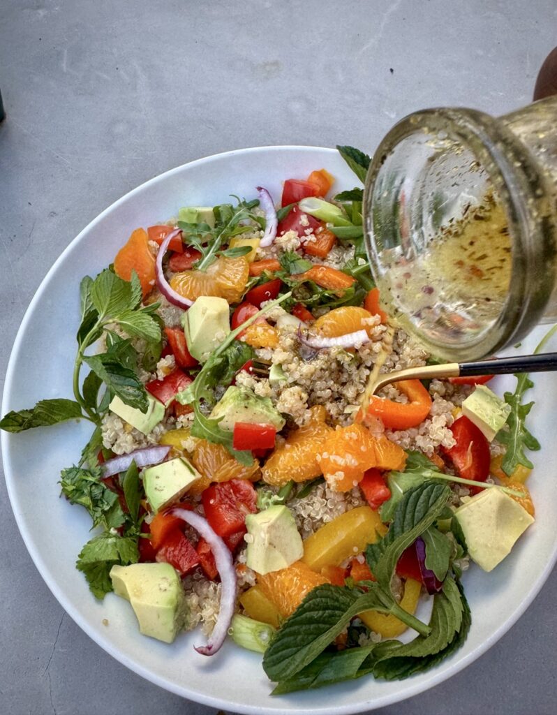 Bowl of Quinoa Citrus Salad with mandarin oranges, bell peppers, avocados, mint, arugula, scallion and arugula  with citrus ginger dressing being poured over it.