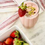 glass of strawberry Lassi garnished with a whole strawberry on a marble countertop with a container of fresh strawberries to the side.