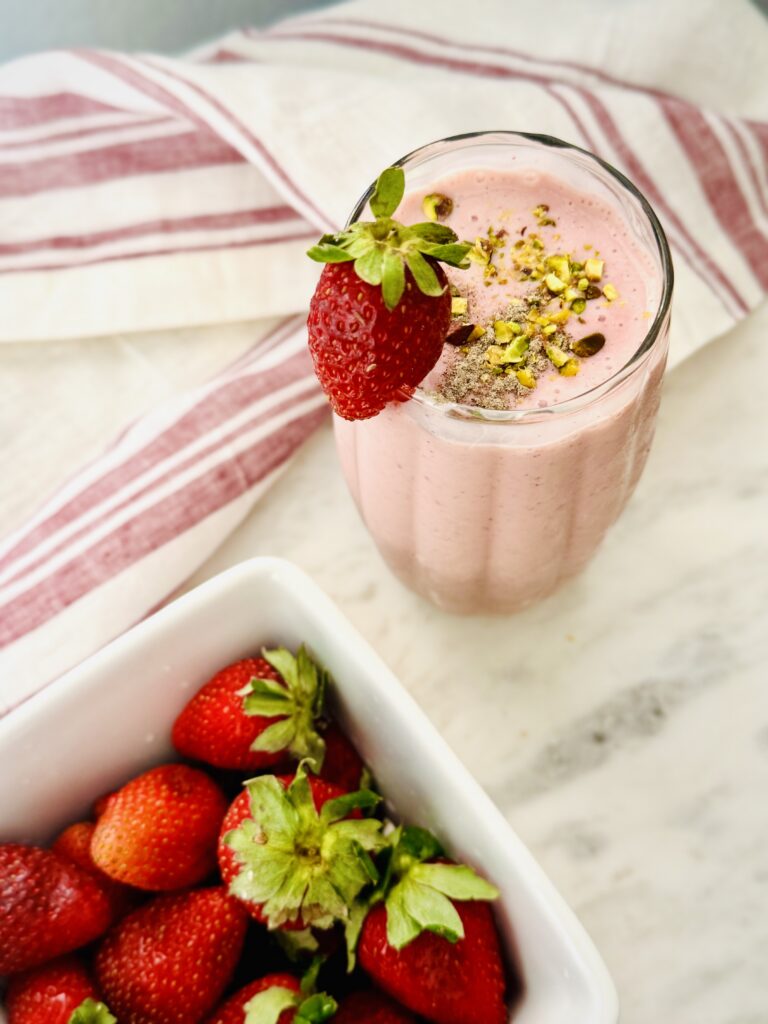 glass of strawberry Lassi garnished with a whole strawberry on a marble countertop with a container of fresh strawberries to the side.