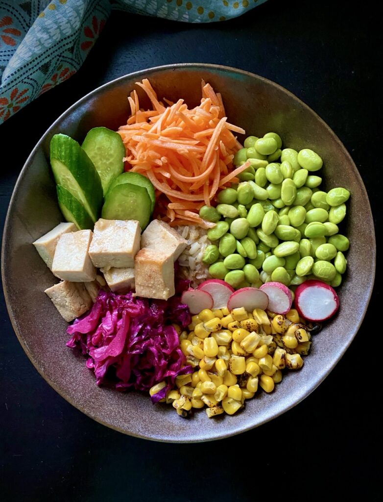 Overhead view of miso bowl with shredded carrots, slices of radish, roasted corn, edamame, pickled cabbage, cubed tofu, and sliced Persian cucumbers over rice.