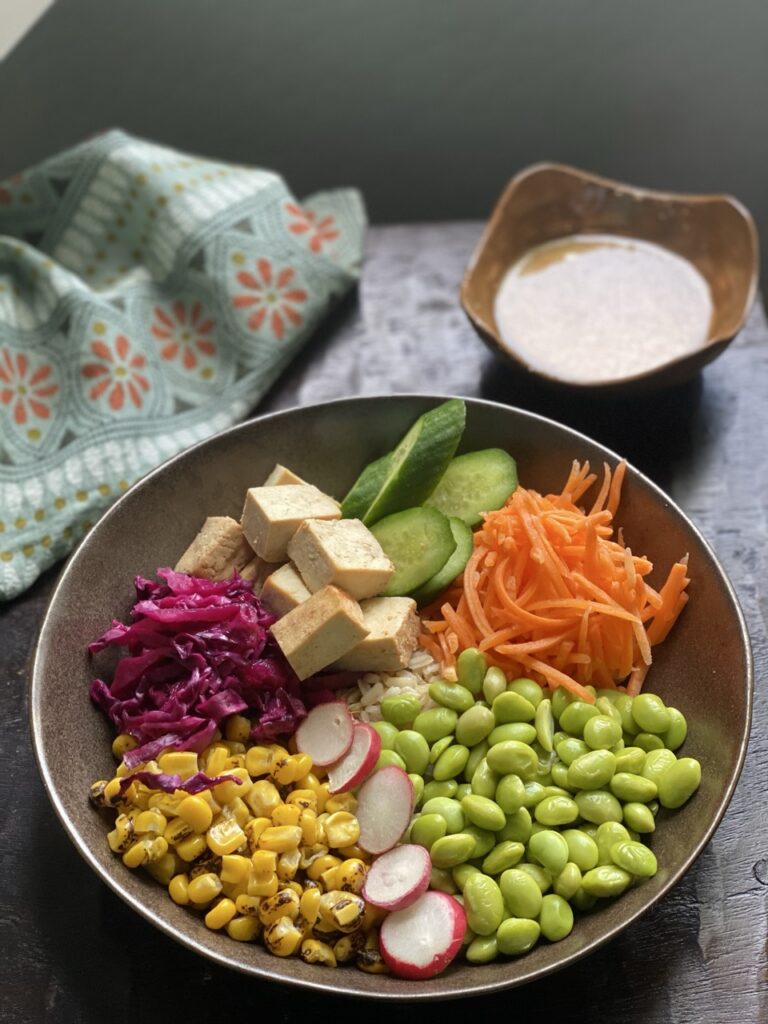 Overhead view of miso bowl with shredded carrots, slices of radish, roasted corn, edamame, pickled cabbage, cubed tofu, and sliced Persian cucumbers over rice with a bowl of lmliso dressing on the side.