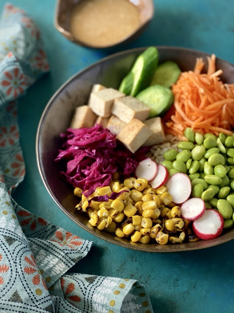 Overhead view of miso bowl with shredded carrots, slices of radish, roasted corn, edamame, pickled cabbage, cubed tofu, and sliced Persian cucumbers over rice.