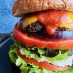 Close up photo of portobello mushroom sandwich on a wheat bun with melted cheese, ketchup, lettuce, tomatoes, and red onion.