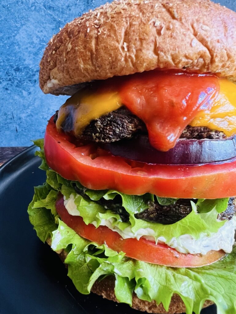 Close up photo of portobello mushroom sandwich on a wheat bun with melted cheese, ketchup, lettuce, tomatoes, and red onion.