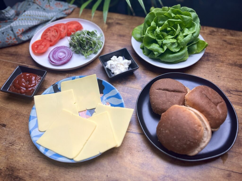 Ingredients for portobello mushroom sandwich displayed in plates on a wooden table. These include 3 wheat buns, 5 slices of vegan cheese, a container of ketchup, a small container of vegan mayo, a plate with sliced tomatoes, a slice of red onion and sprouts, and plate of butter lettuce.