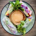 Summer rolls filled with bell peppers, red cabbage, lettuce, vermicelli, radishes, and cucumbers with bowl of peanut sauce.