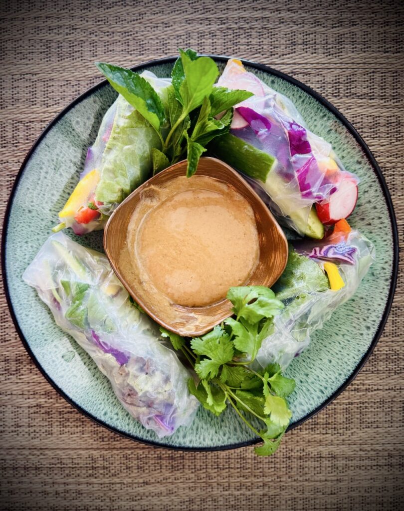 Summer rolls filled with bell peppers, red cabbage, lettuce, vermicelli, radishes, and cucumbers with bowl of peanut sauce.