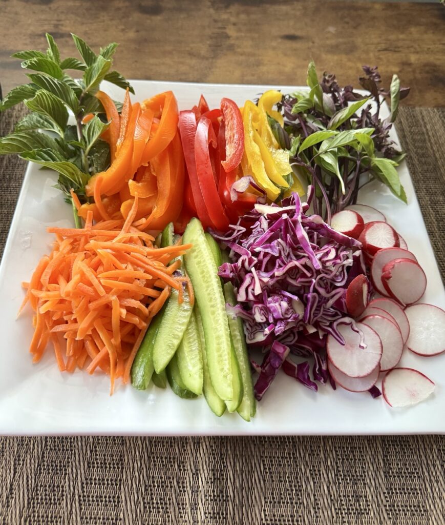 A platter with julienned orange, red and yellow bell peppers, julienned cucumbers, shredded carrots and purple cabbage, sliced radishes and fresh mint and basil.