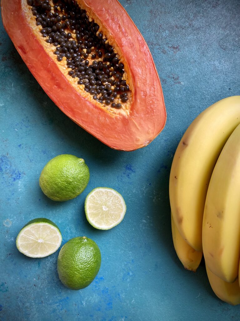 Halved papaya, bananas, and limes on a blue-green background.