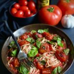 Bowl of spaghetti with fresh tomato sauce topped with charred tomatoes and fresh basil. Vine ripened tomatoes, a bulb of garlic, a bottle of olive oil, and a bowl of cherry tomatoes are in the background.