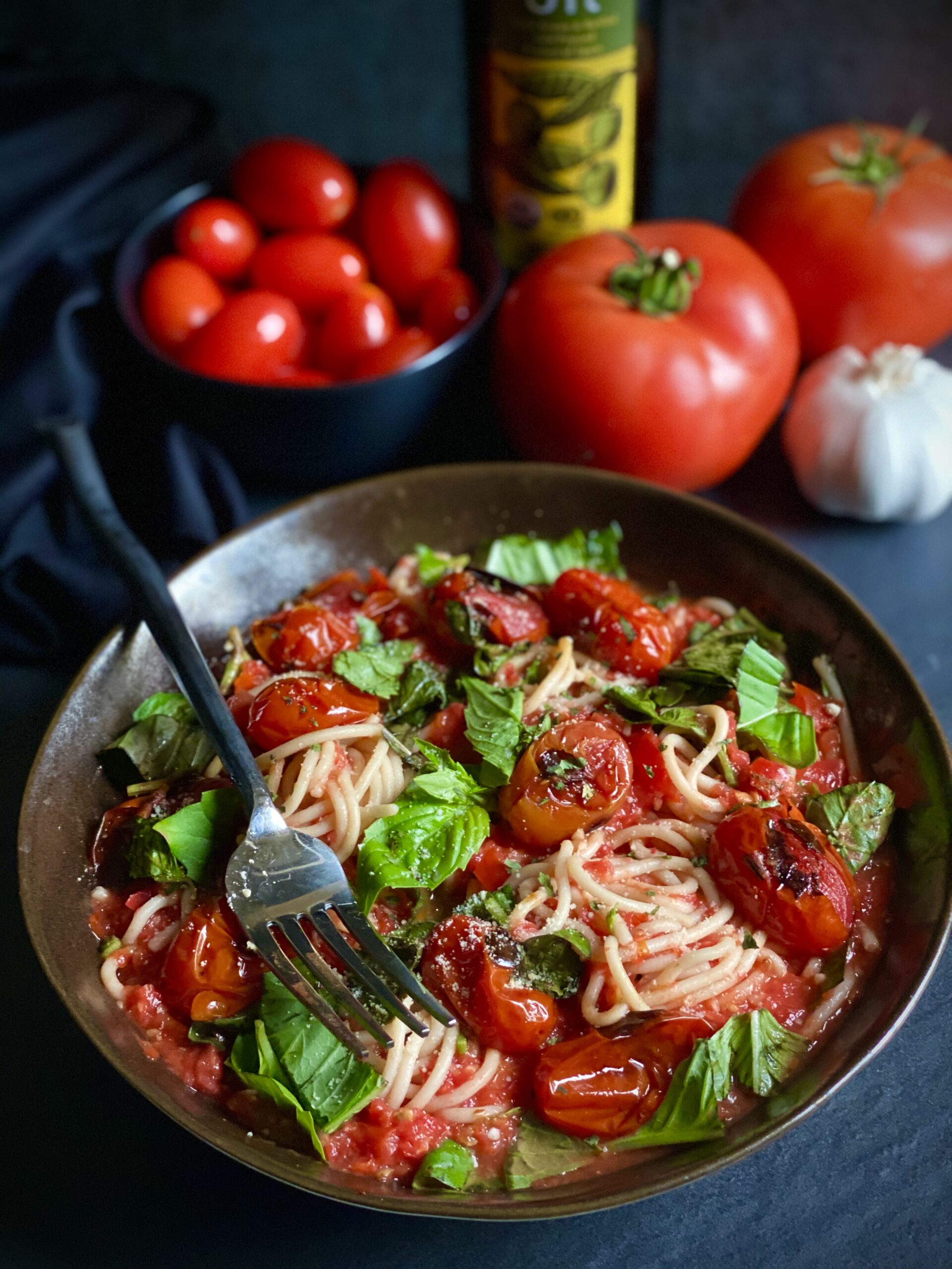 Bowl of spaghetti with fresh tomato sauce topped with charred tomatoes and fresh basil. Vine ripened tomatoes, a bulb of garlic, a bottle of olive oil, and a bowl of cherry tomatoes are in the background.