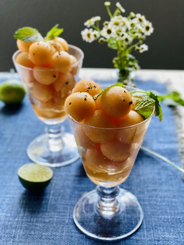 Two footed glasses filled with balls of cantaloupe garnished with mint with a vase of flowers in the background and lime halves it the foreground.