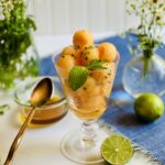 Footed glass filled with balls of cantaloupe garnished with mint with a small bowl of honey and vase of flowers in the background and lime halves it the foreground.