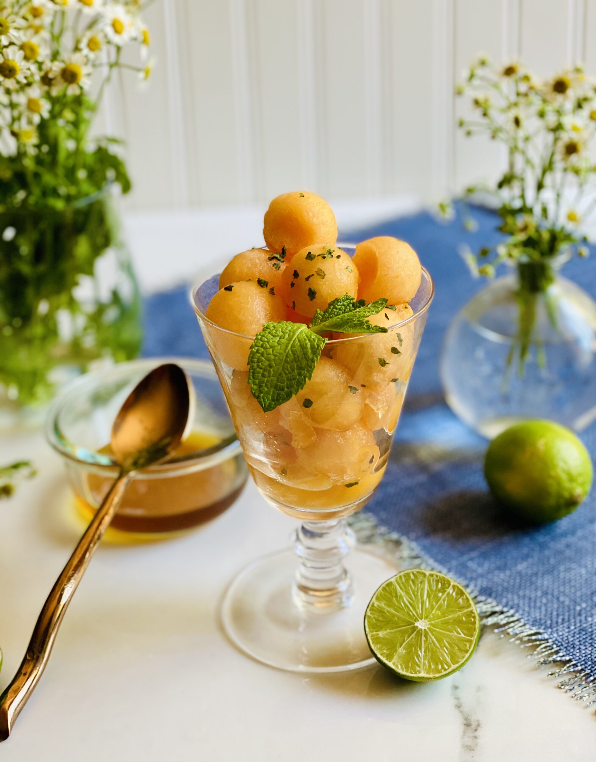 Footed glass filled with balls of cantaloupe garnished with mint with a small bowl of honey and vase of flowers in the background and lime halves it the foreground.