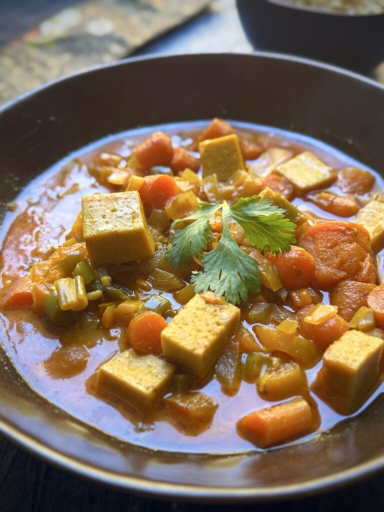 Bowl of vegan panang curry with cubes of tofu, bell peppers, carrots, sweet potato, coconut milk, spring onion and cilantro