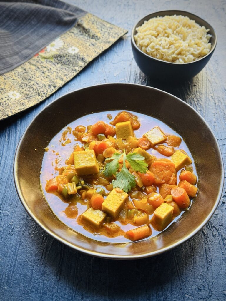 Bowl of vegan panang curry with cubes of tofu, bell peppers, carrots, sweet potato, coconut milk, spring onion and cilantro with a smaller bowl of brown jasmine rice on the side.
