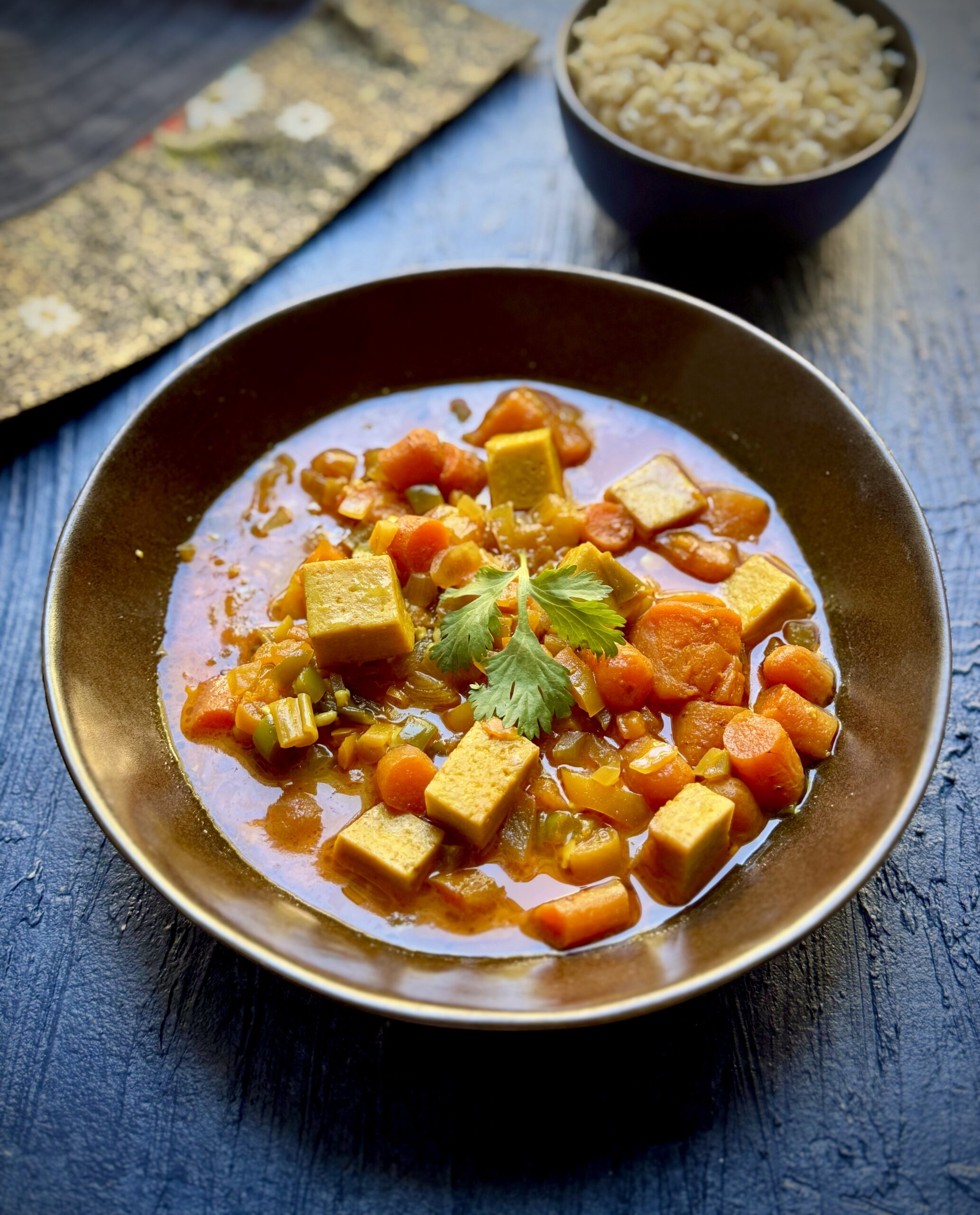 Bowl of vegan panang curry with cubes of tofu, bell peppers, carrots, sweet potato, coconut milk, spring onion and cilantro