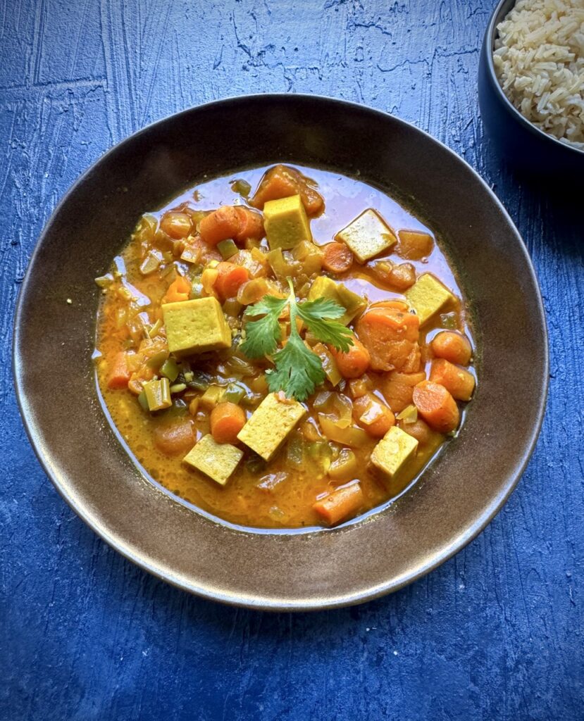 Bowl of vegan panang curry with cubes of tofu, bell peppers, carrots, sweet potato, coconut milk, spring onion and cilantro