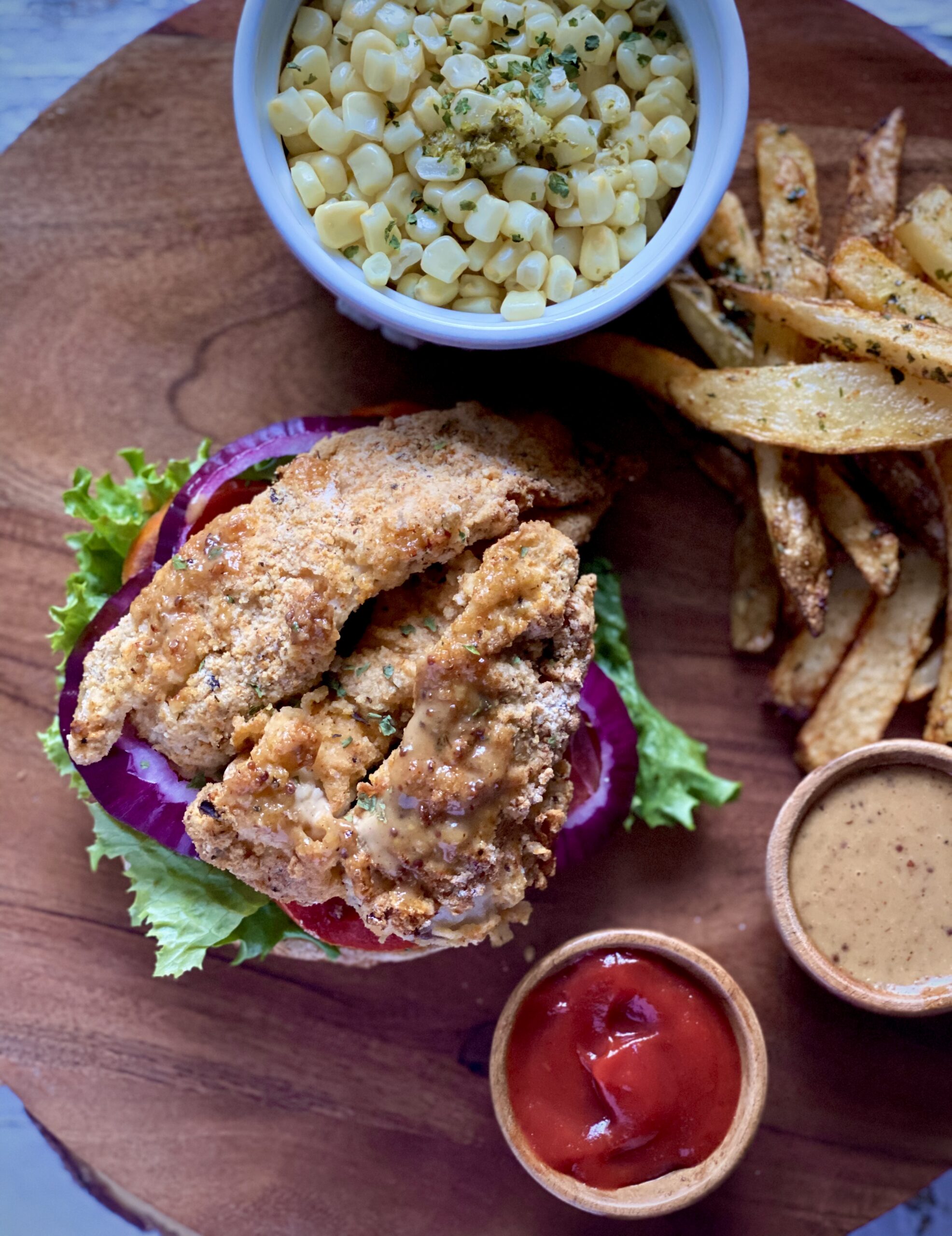 Open-faced oyster mushroom sandwich with lettuce and tomatoes, a side of oven fries, with a container of corn and another with sauce and with ketchup.