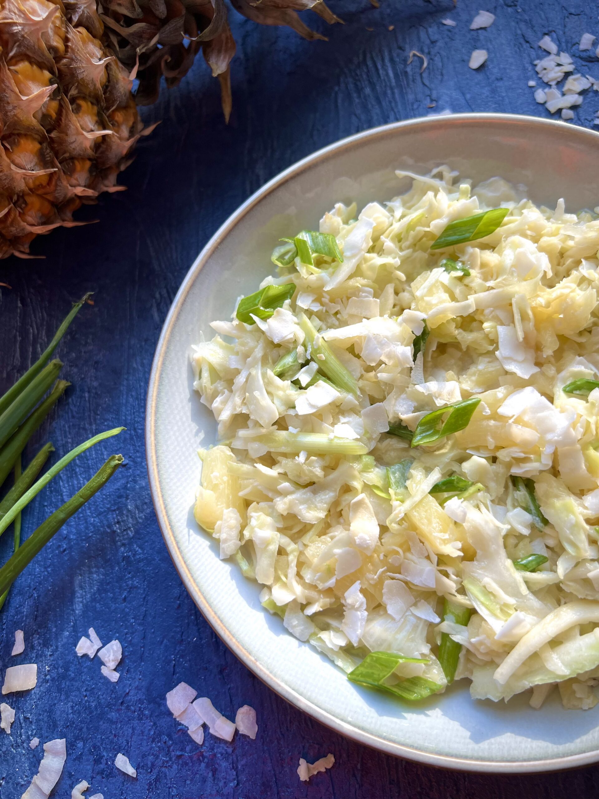 Bowl of tropical coleslaw with shredded coconut, crushed pineapple, and scallions with a fresh pineapple in the background.