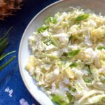 Bowl of tropical coleslaw with shredded coconut, crushed pineapple, and scallions with a fresh pineapple in the background.
